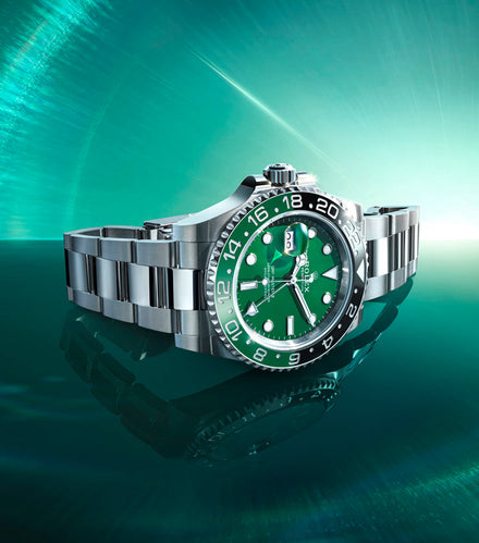 A stainless steel luxury watch with a green dial and bezel displayed on a reflective teal surface.